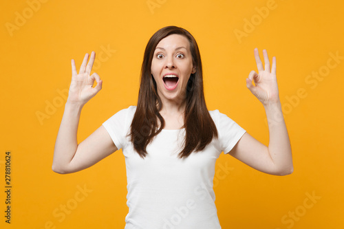 Portrait of amazed young woman in white casual clothes keeping mouth open, showing OK gesture isolated on bright yellow orange wall background in studio. People lifestyle concept. Mock up copy space.