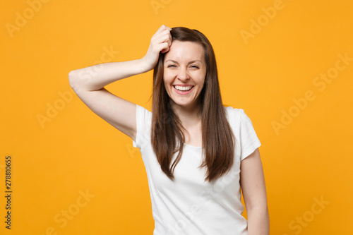 Portrait of laughing young woman in white casual clothes looking camera, putting hand on head isolated on bright yellow orange wall background in studio. People lifestyle concept. Mock up copy space.