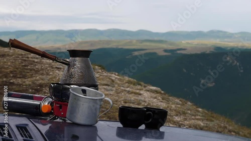 metal cezve with wooden handle stands on modern gas burner near cups against fantastic mountains closeup slow motion photo