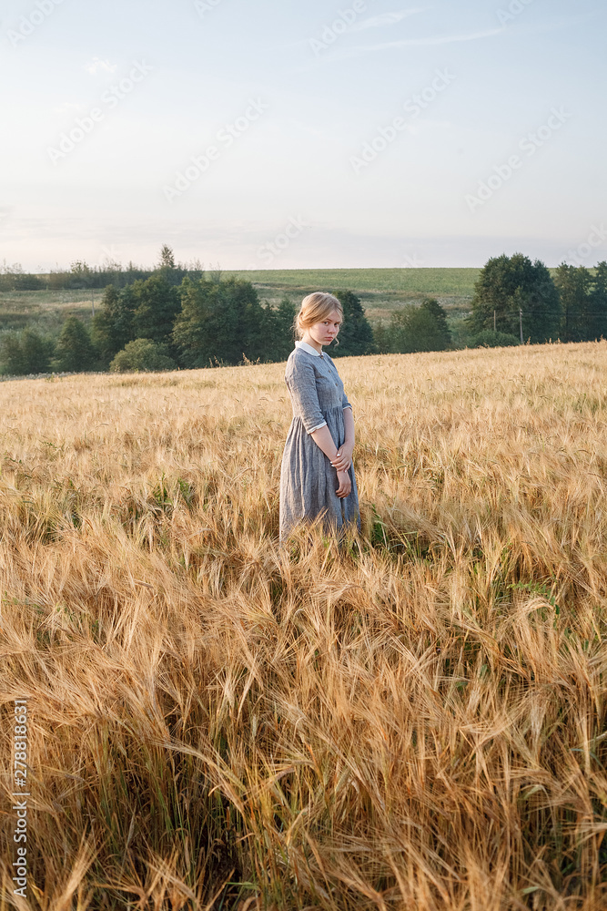 young girl in blue dress with collected hair looks at camera in field of ears at sunrise. Green boscage in background young girl in blue dress with collected hair looks at camera in field of ears at sunrise. Green boscage in background