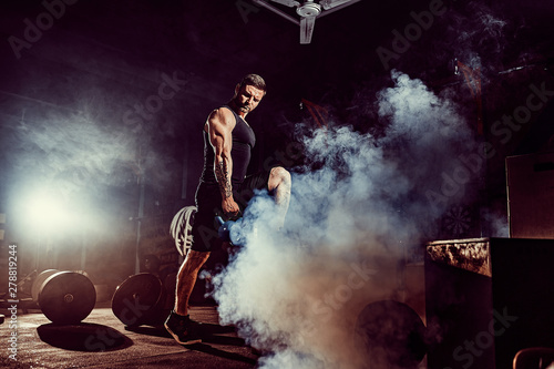 Muscular attractive caucasian bearded tasttoed man lifting kettlebell in a gym. Weight plates  dumbbell and tires in smoke background.