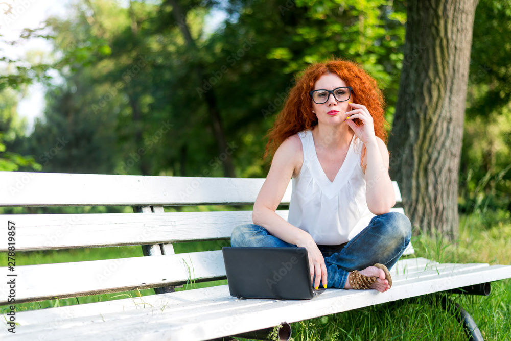 Red-haired female freelancer working on a laptop while sitting on a park bench.