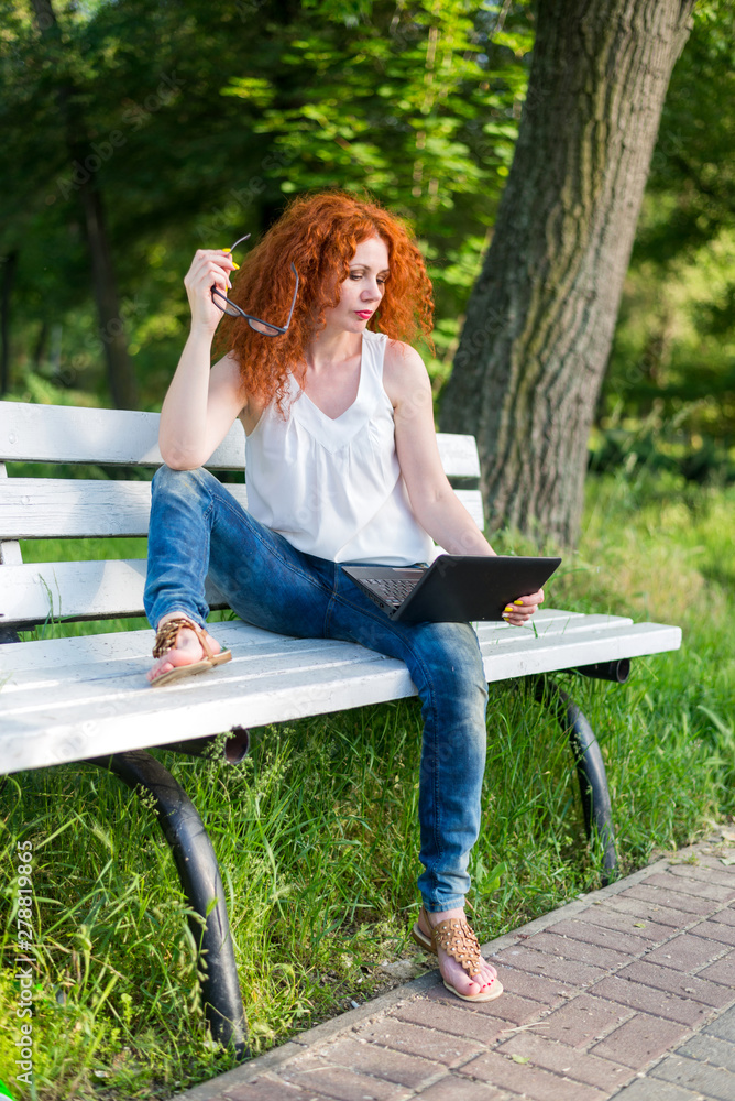 Naklejka premium Red-haired female freelancer working on a laptop while sitting on a park bench.