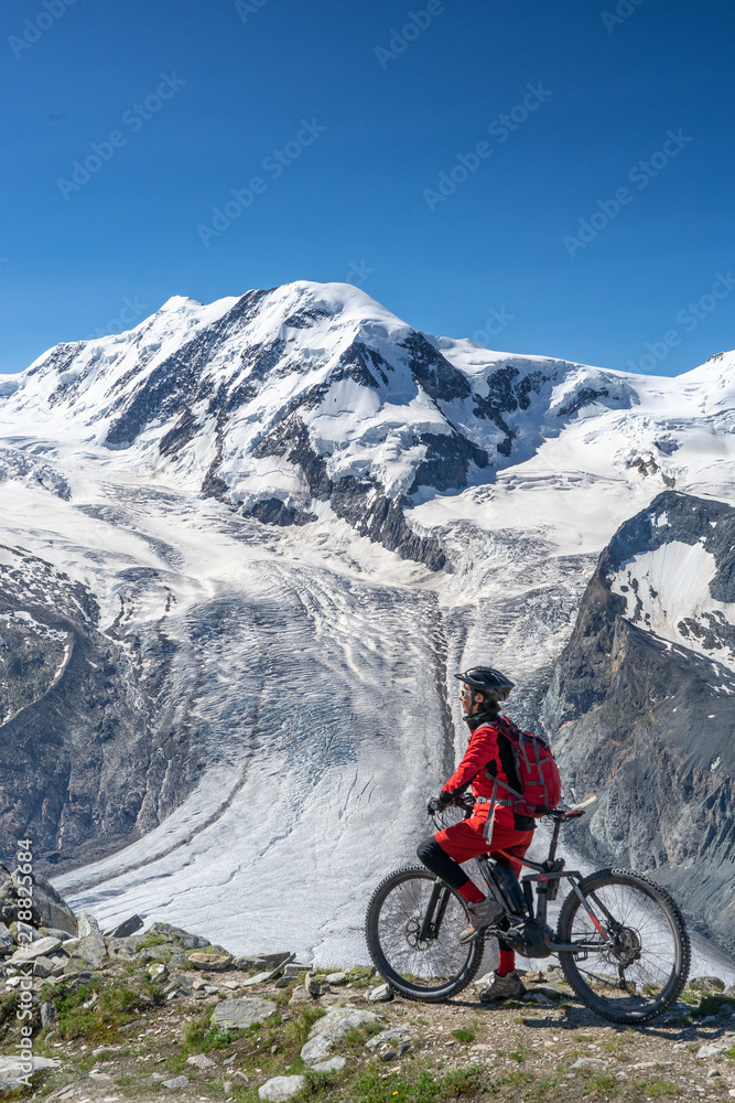 active senior woman, riding her electric mountainbikeon the Gornergrat in Zermatt, Wallis,Switzerland. In The background Gorner Glacier, Monte Rosa, Liskam am Breithorn
