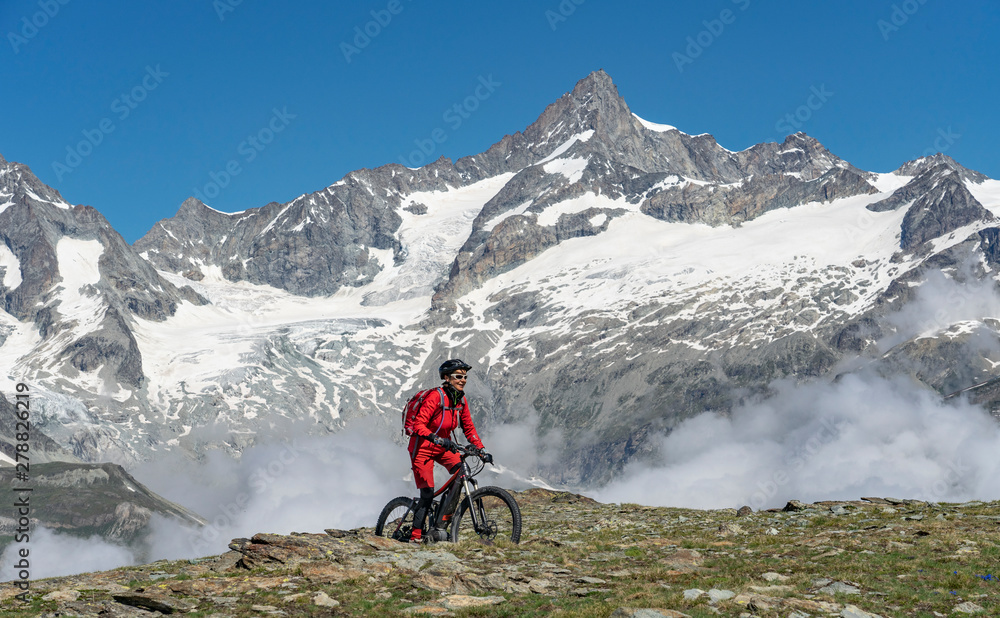 active senior woman, riding her electric mountainbikeon the Gornergrat in Zermatt, Wallis,Switzerland. In The background Weisshorn, Zinalrothorn and Obergabelhorn