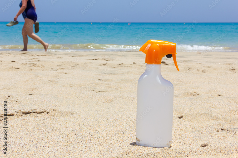 Sunblock white bottle on sand as woman passes by shore on turquoise water beach. Sunscreen spray, skin care protection, summer vacation concepts Sunblock white bottle on sand as woman passes by shore on turquoise water beach. Sunscreen spray, skin care protection, summer vacation concepts