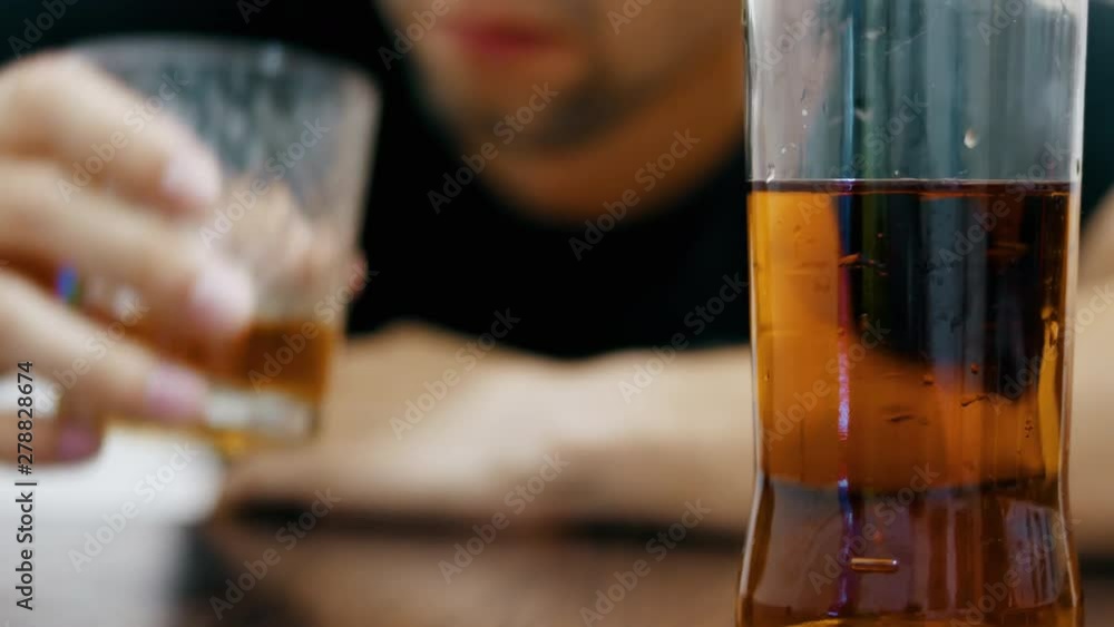 In the background a blurred young drunk man drinks whiskey from a glass and falls asleep on the table, a half-empty bottle of whiskey in the foreground in focus In the background a blurred young drunk man drinks whiskey from a glass and falls asleep on the table, a half-empty bottle of whiskey in the foreground in focus