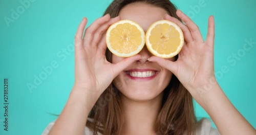 Funny portrait of young attractive brunette on turquoise backgroung is taking splitted halfs of yellow lemon and closes his eyes, smiling and laughing. Slow motion.
