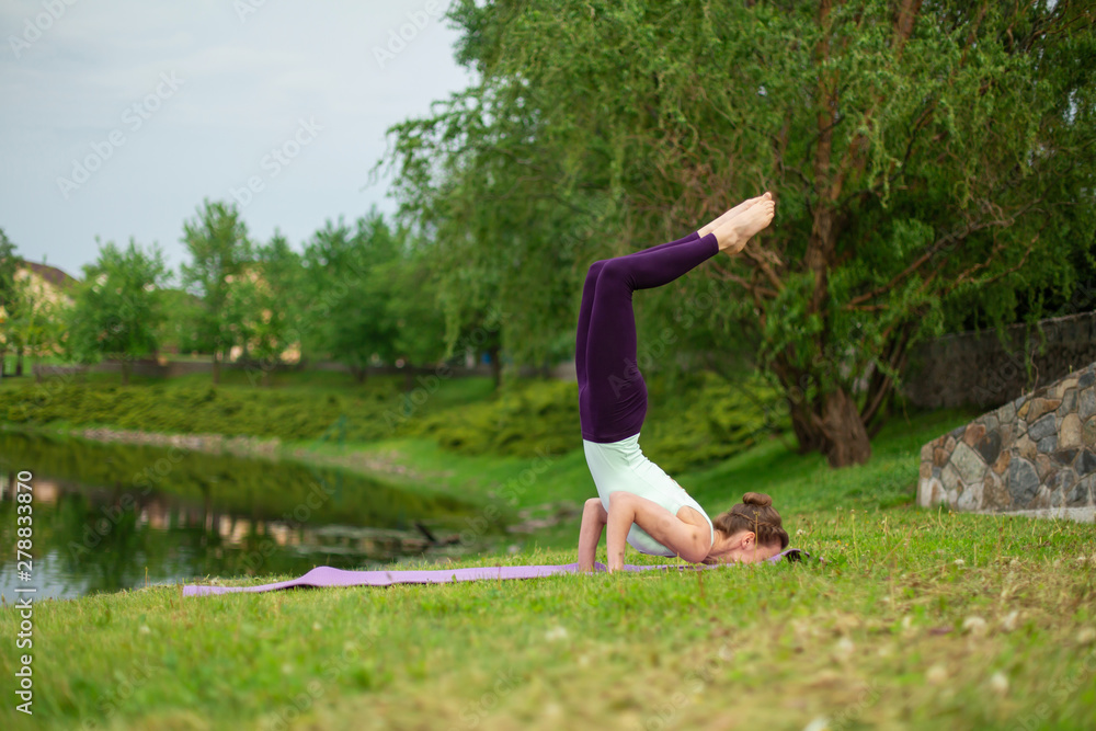 Slender young brunette yogi performs complex yoga exercise on green grass in the summer on a background of nature Slender young brunette yogi performs complex yoga exercise on green grass in the summer on a background of nature
