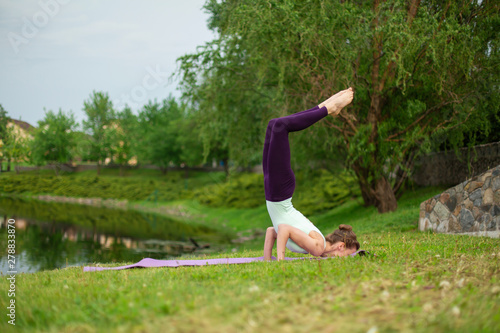 Slender young brunette yogi performs complex yoga exercise on green grass in the summer on a background of nature