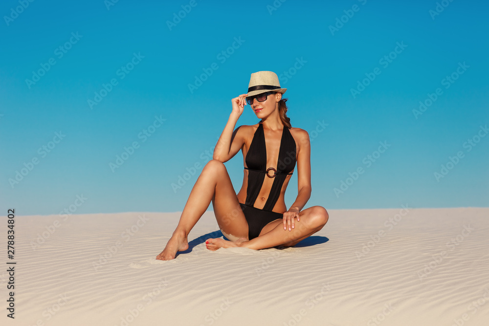 Portrait of sexy beautiful tanned model woman posing in fashion black swimwear bikini, fedora hat and sunglasses sand desert beach. Exotic country travel and rest concept. Slim figure and sport body. - obrazy, fototapety, plakaty 