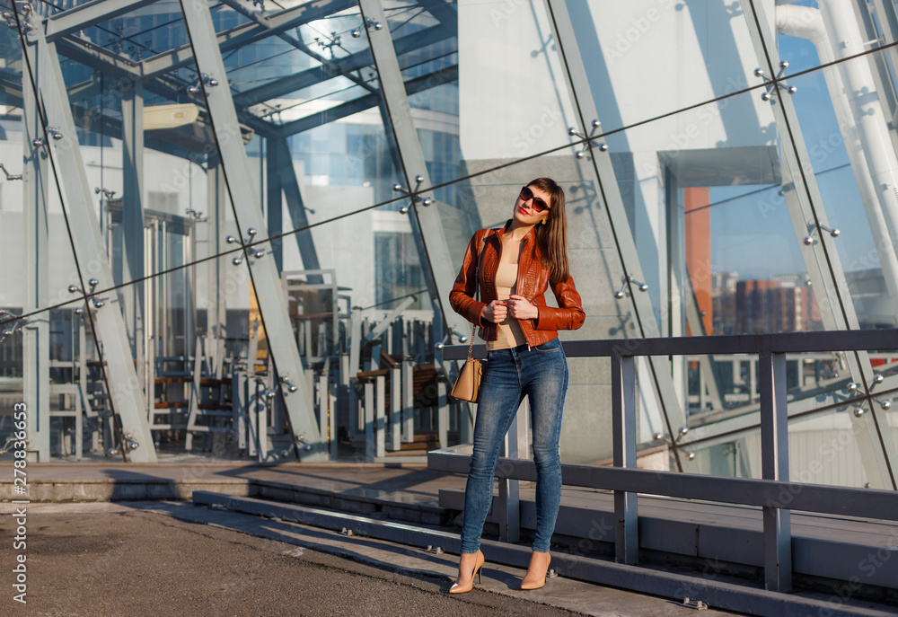 Portrait of beautiful brunette young woman in nice red brown jacket, denim jeans and sunglasses. Genuine leather bag, high heels shoes. Spring autumn fashion trendy photo of lady on urban background.