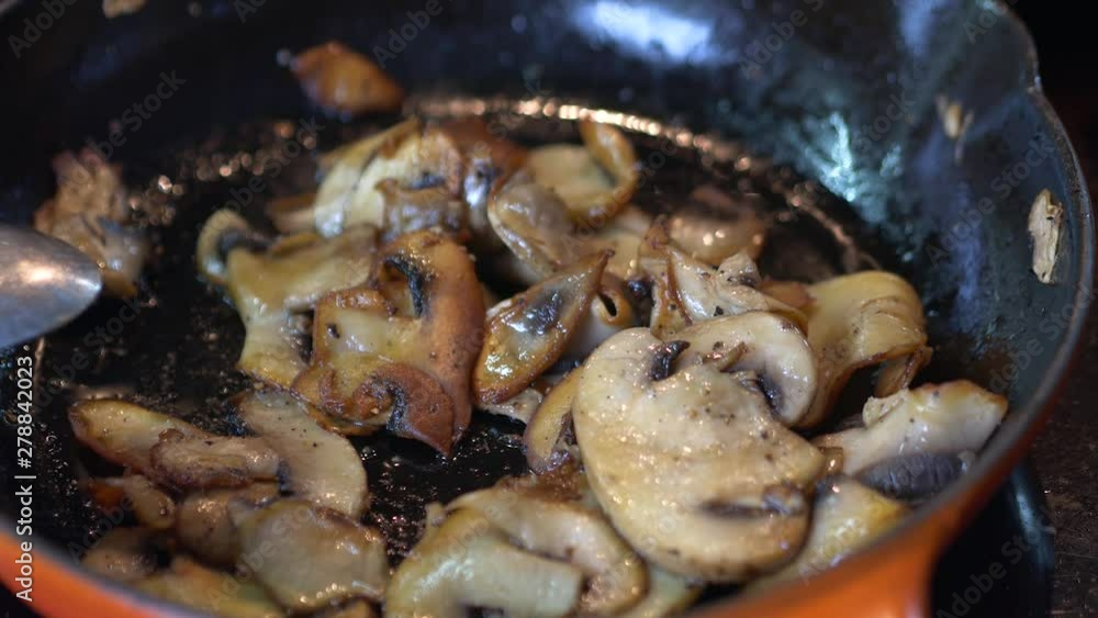 Sliced mushrooms are frying in a hot red pan. Occasionally, the mushrooms are stirred by a spoon. Salt and pepper is added. Sliced mushrooms are frying in a hot red pan. Occasionally, the mushrooms are stirred by a spoon. Salt and pepper is added.