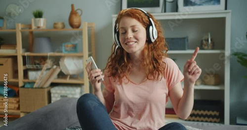 Joyful young lady is enjoying music in headphones using smartphone dancing sitting on bed in apartment. House, devices and happy carefree people concept. photo