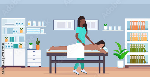 girl having back massage african american masseuse in uniform massaging patient body woman relaxing lying on bed treatments concept modern hospital office interior full length horizontal