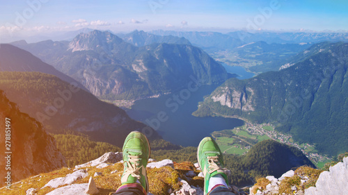 Legs of traveler in stylish green sneakers sitting on a high mountain cliff enjoying scenery mountain top. Pov view Hiking freedom concept. Austria Hallstatter See lake Krippenstein mountain Hallstatt