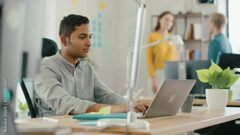 Smart and Handsome Indian Office Worker Sitting at His Desk works on a Laptop. In the Background Modern Office with Diverse Team of Young Professionals Working Smart and Handsome Indian Office Worker Sitting at His Desk works on a Laptop. In the Background Modern Office with Diverse Team of Young Professionals Working