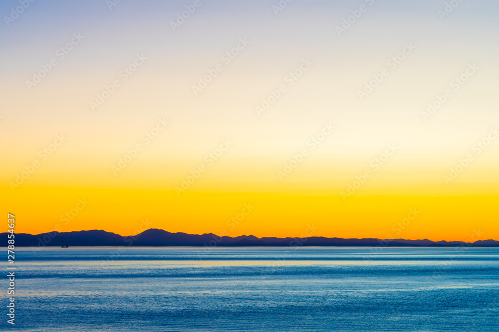 Obraz premium Beautiful twilight with golden sunset/sunrise over the ocean behind mountain silhouette. Orange yellow blue skies over water as the sun sets/rises. Harmony and beauty in nature. Scenic peaceful view.