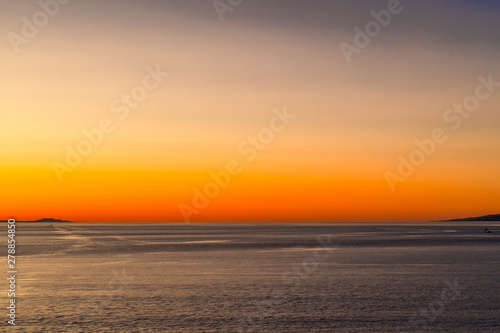 Colorful golden orange yellow sunset/sunrise twilight behind mountain silhouette over the calm ocean water. Scenic peaceful landscape seascape nature setting as the sun sets/rises beyond the calm seas