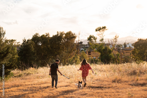 Back view of of cheerful couple running with small friendly dog among yellow grass in park