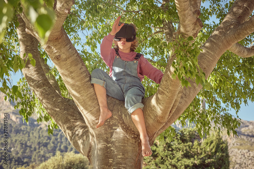 Enthusiastic boy in virtual reality glasses and denim overalls sitting high on tree in bright day