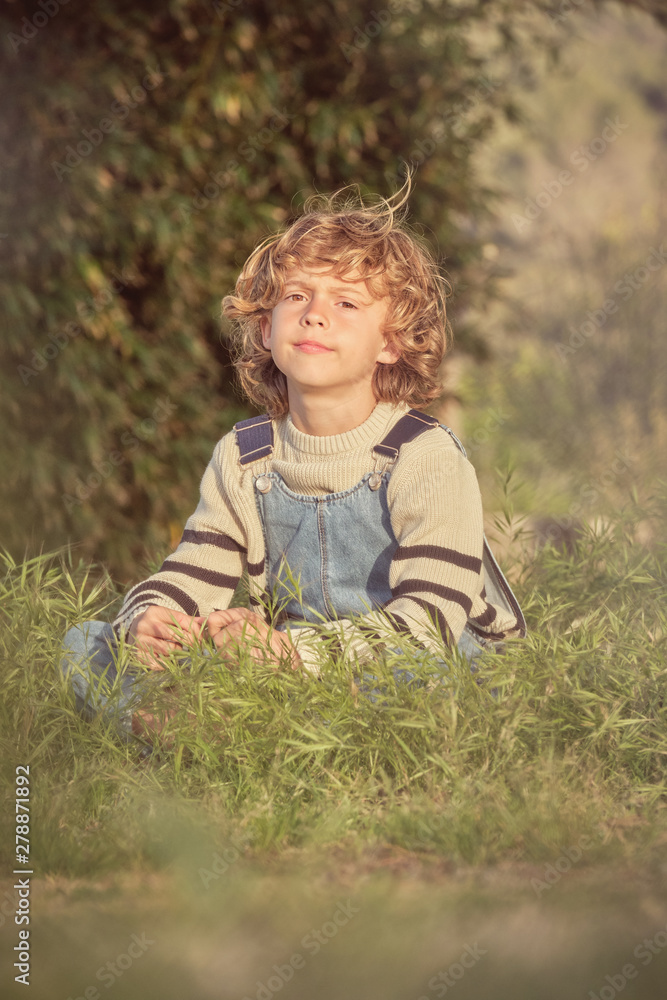 Adorable serious child in denim overalls sitting in green grass and looking at camera in sunny day Adorable serious child in denim overalls sitting in green grass and looking at camera in sunny day