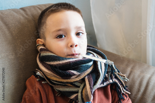 Young boy sitting on the sofa. Ill child waiting doctor to visit him. Influenza, pediatric service at home. Medical concept of seasonal illnesses. photo