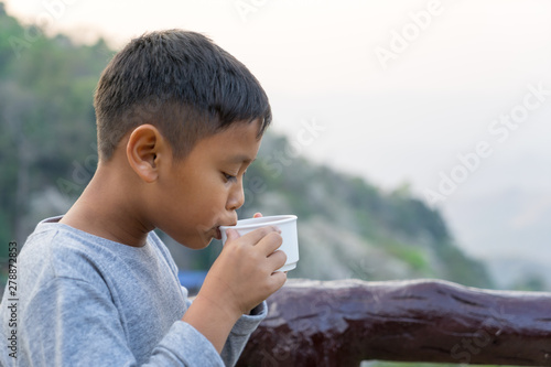 Asian kid boy is drinking water from the glass. With mountain views background