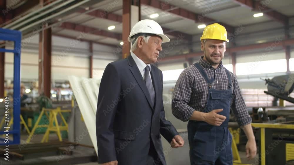 Tracking pan shot of smiling factory engineer and mature executive walking along workshop, engaged in lively discussion, then stopping to look at some objects at distance, with pointing and gesturing Tracking pan shot of smiling factory engineer and mature executive walking along workshop, engaged in lively discussion, then stopping to look at some objects at distance, with pointing and gesturing