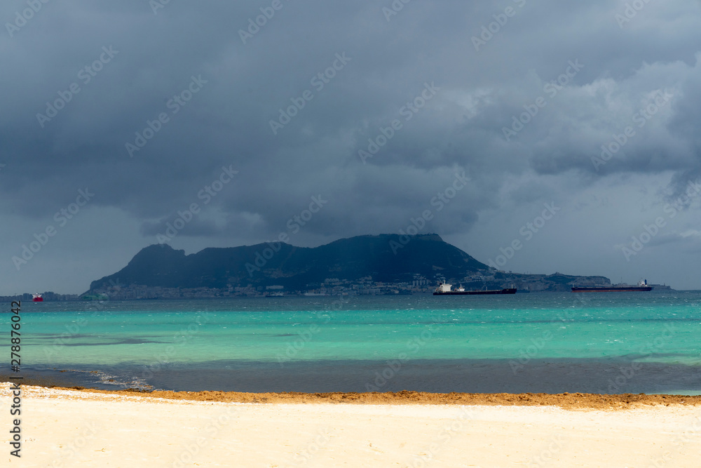 Colorful view from the beach de Getares: white sand contrast with azure sea and dark sky. Fluffy clouds over the Gibraltar. Impressive ships are constantly in the Bay. Algeciras, Andalusia, Spain.