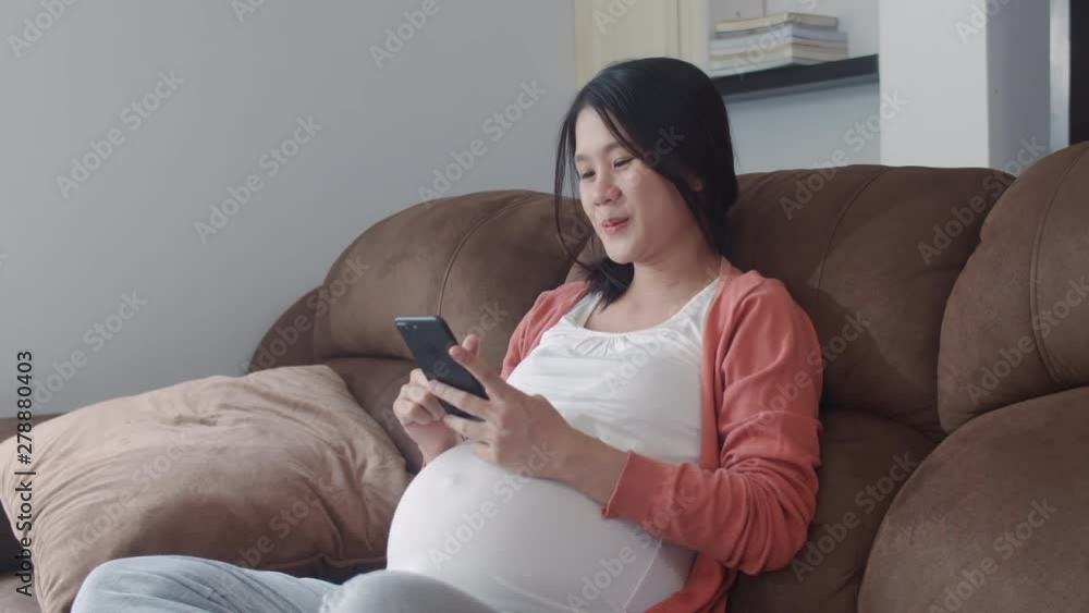 Young Asian Pregnant woman using mobile phone search pregnancy information. Mom feeling happy smiling positive and peaceful while take care her child lying on sofa in living room at home concept. Young Asian Pregnant woman using mobile phone search pregnancy information. Mom feeling happy smiling positive and peaceful while take care her child lying on sofa in living room at home concept.