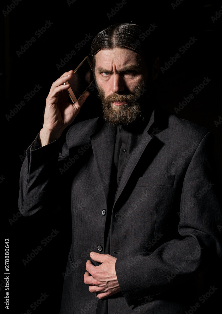 strange bearded senior priest with a smartphone, bearded old man is calling on a dark background