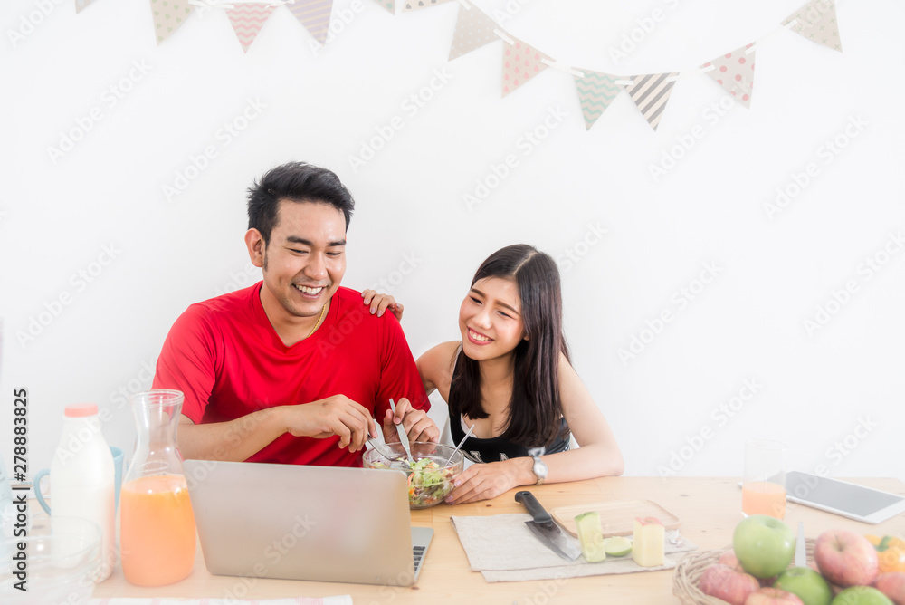 Happy Asian couple using laptop and eating salad and fresh fruit at home.