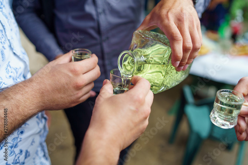 Close up on midsection of male hands holding drinking glasses pouring homemade domestic brandy slivovitza standing outdoors celebrating photo