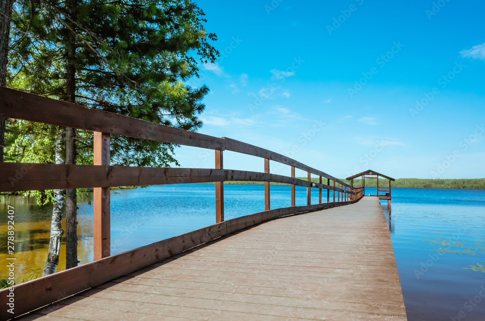 Pier on the lake with calm water under a bright blue sky. Soothing calm minimalistic landscape. Trees surround the pond. Place for text.