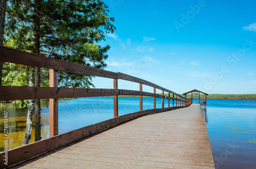 Pier on the lake with calm water under a bright blue sky. Soothing calm minimalistic landscape. Trees surround the pond. Place for text. © Ольга Холявина