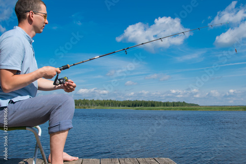 a man in yellow glasses, sitting on the pier, holding a spinning, fishing, against a beautiful landscape