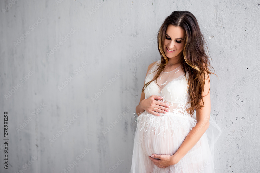 beautiful pregnant woman in white lace dress posing over gray wall background with copy space beautiful pregnant woman in white lace dress posing over gray wall background with copy space