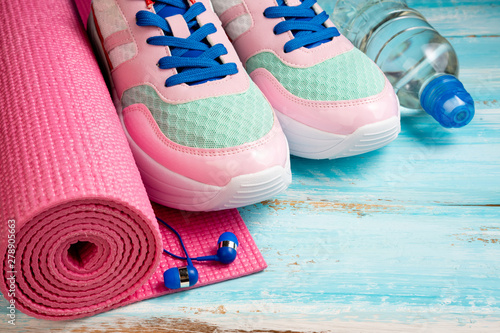 Pink yoga mat, sport shoes, bottle of water and earphones on blue wooden background. Sport, healthy lifestyle, yoga concept. Trendy female sport equipment. Copy space