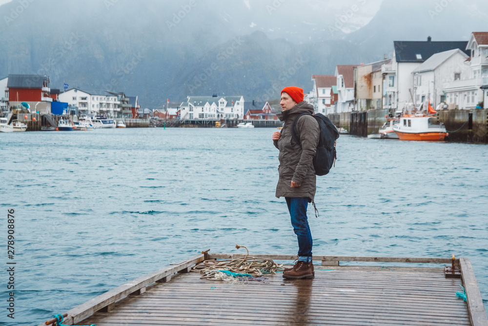 Traveler man with a backpack wearing a red hat standing on the background of fishing houses, ships, mountain and lake wooden pier. Travel lifestyle concept.