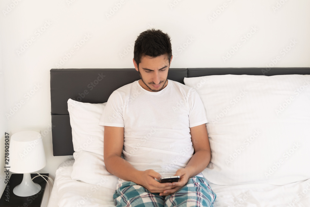 Young handsome man sitting on the bed. He uses a mobile phone. The problem of dependence on mobile social networks. Young handsome man sitting on the bed. He uses a mobile phone. The problem of dependence on mobile social networks.