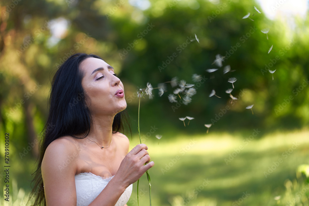 Young pretty brunette woman in white wedding dress blowing on dandelion Young pretty brunette woman in white wedding dress blowing on dandelion