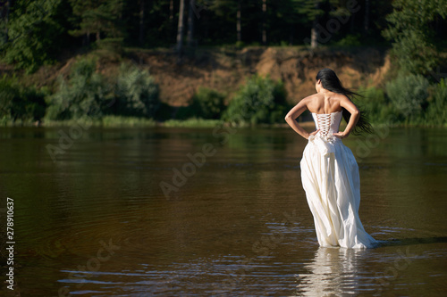 Young pretty brunette woman in white wedding dress walking on the river barefoot