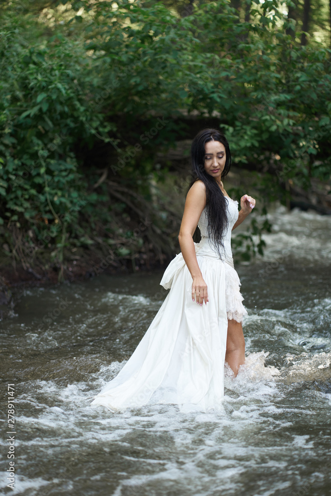 Young pretty brunette woman in white wedding dress stands outdoors in rushing river