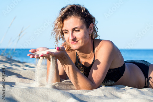 tanned young attractive woman with curly hair in black swimsuit on the beach, keeping sand in palms and sifting it through her fingers
