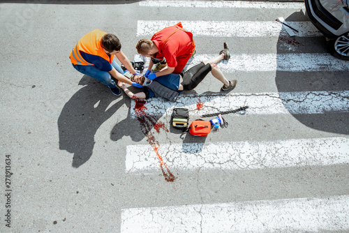 Ambulance worker with man in road vest applying emergency medical care to the injured bleeding person lying on the pedestrian crossing after the accident, view from the above