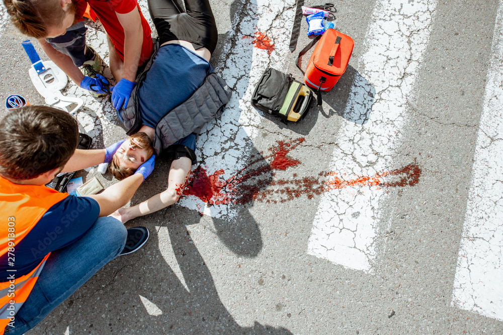 Ambulance worker with man in road vest applying emergency medical care to the injured bleeding person lying on the pedestrian crossing after the accident, view from the above