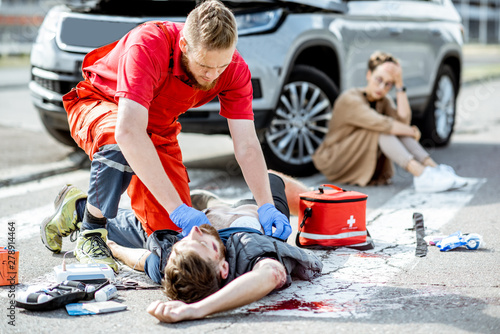 Ambluence worker applying emergency care to the injured bleeding man lying on the pedestrian crossing, dispair woman driver sitting on the background photo