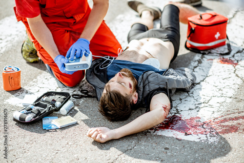 Ambluence worker applying emergency care with defibrillator to the injured bleeding man lying on the pedestrian crossing photo