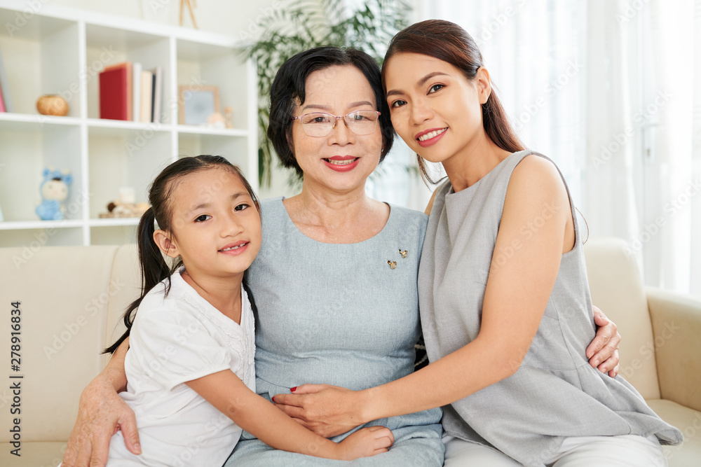 Portrait of happy Asian family generation mother daughter and grandmother embracing and smiling at camera while resting on couch Portrait of happy Asian family generation mother daughter and grandmother embracing and smiling at camera while resting on couch